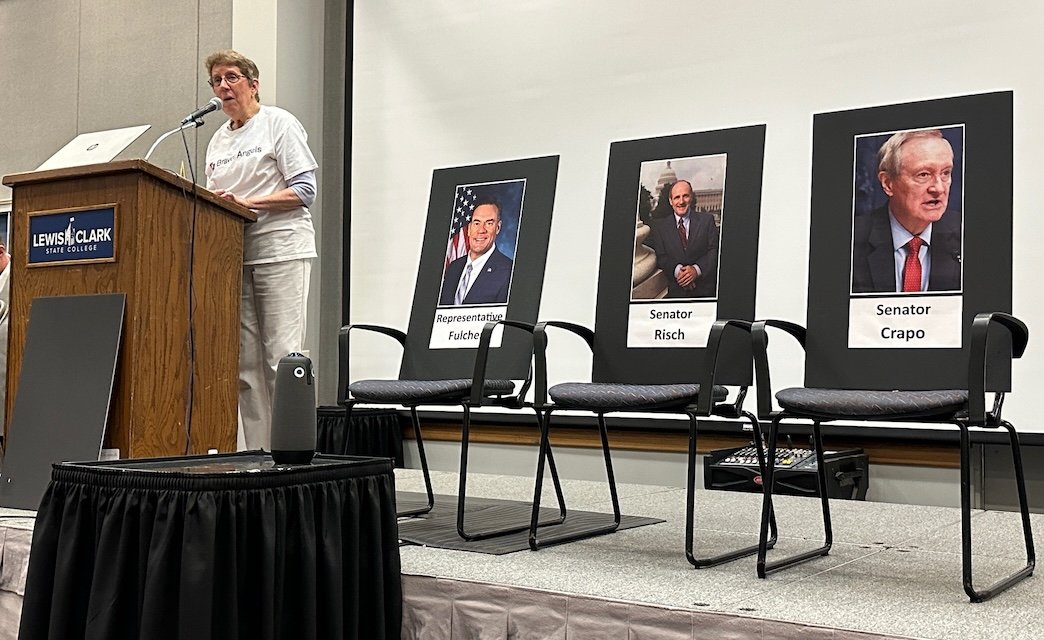 Kathy Dawes speaks to the audience at the Forum for Our Members of Congress on August 23, 2025. Beside her are three chairs with photos of Congressman Fulcher and Senators Risch and Crapo, none of whom attended the Forum.